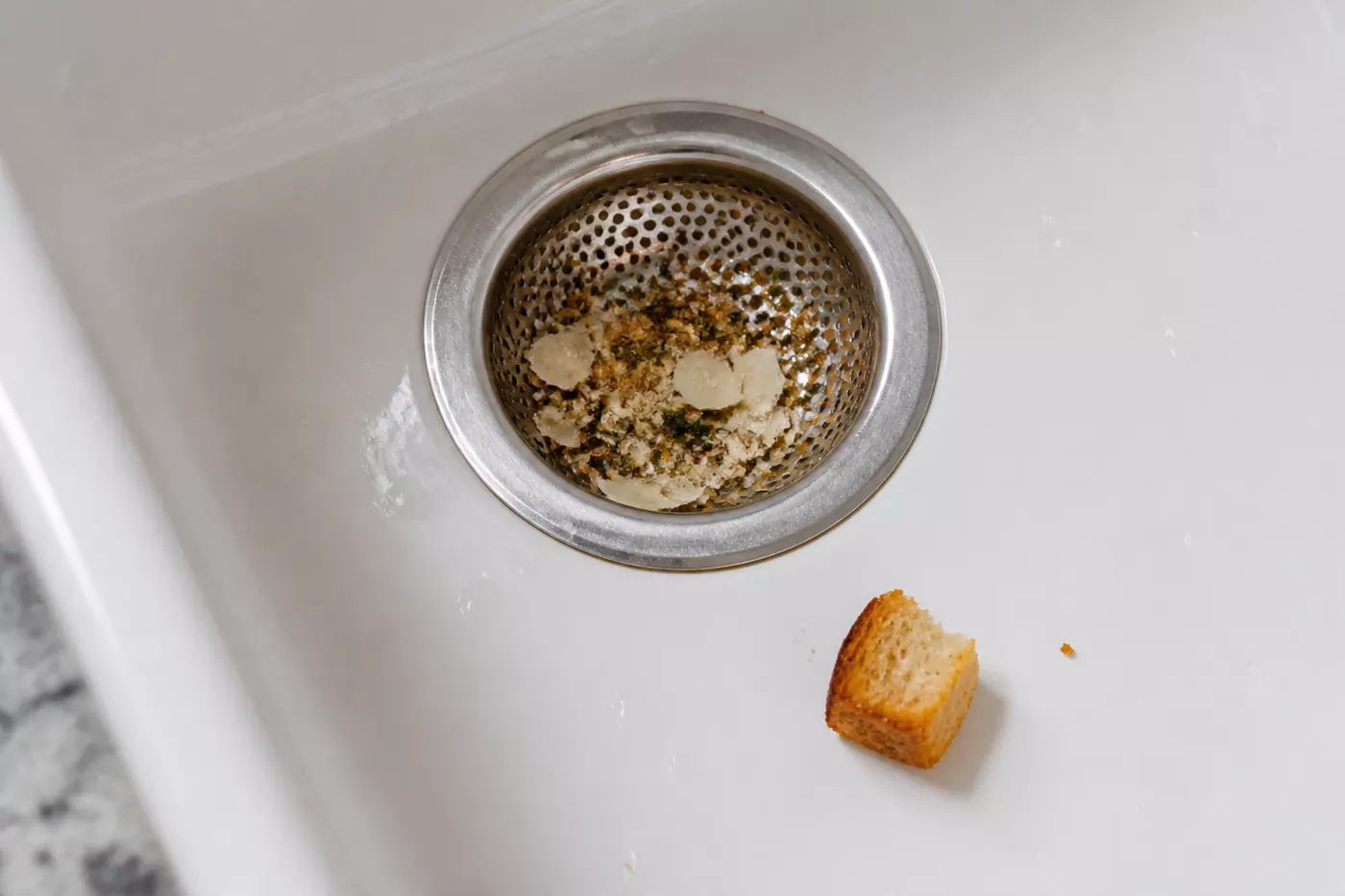 Herb crumbs and Parmesan caught in a steel strainer beside a golden crouton in a white ceramic sink