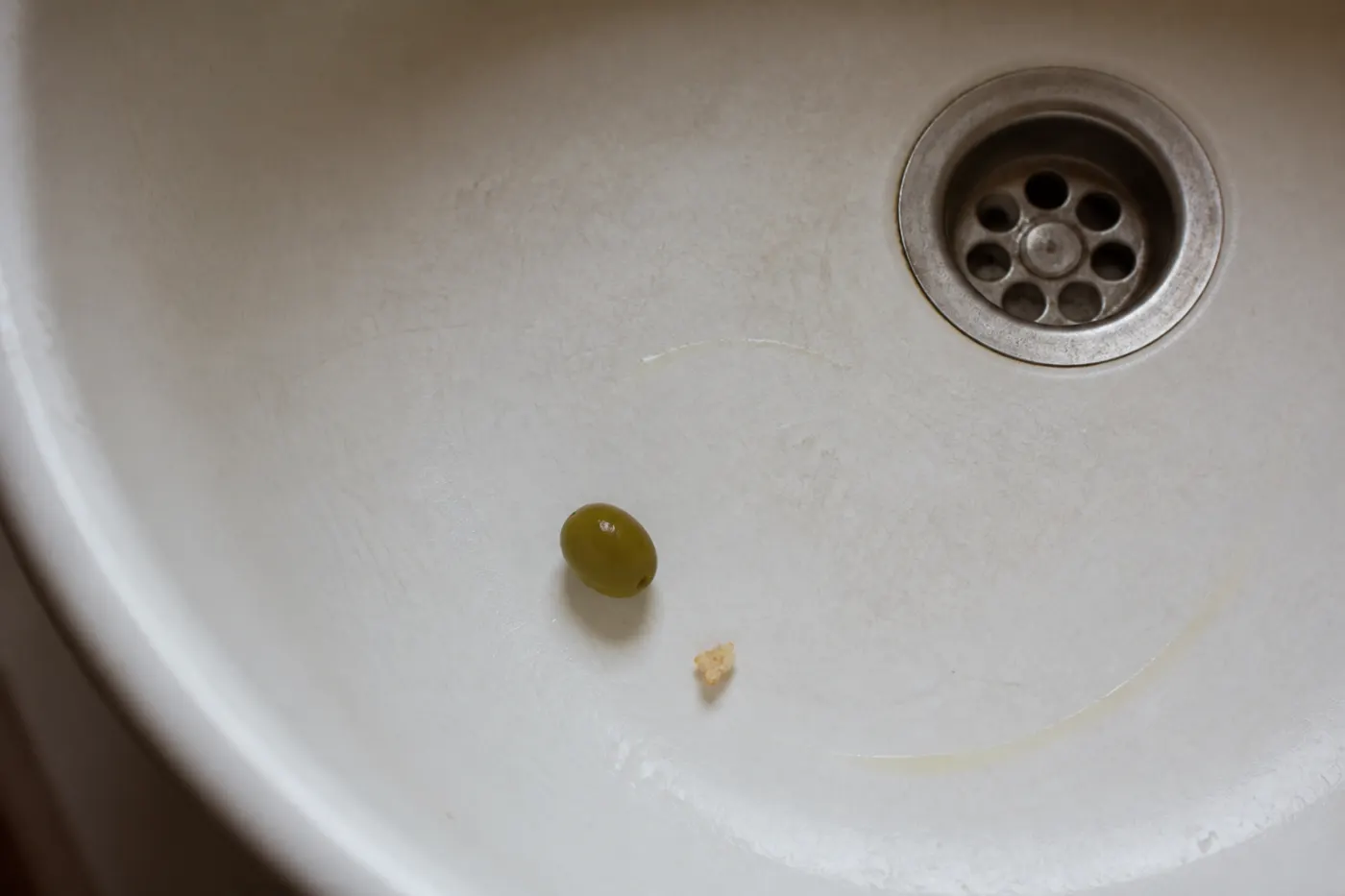 Single green olive, bread crumb and faint oil trace in an old white porcelain sink