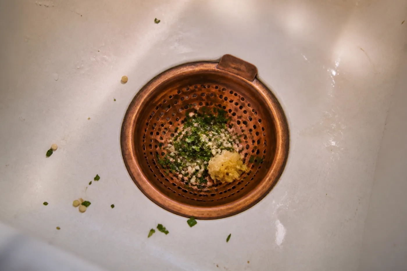 Copper-toned strainer in a white farmhouse sink catching parsley, couscous and lemon pulp