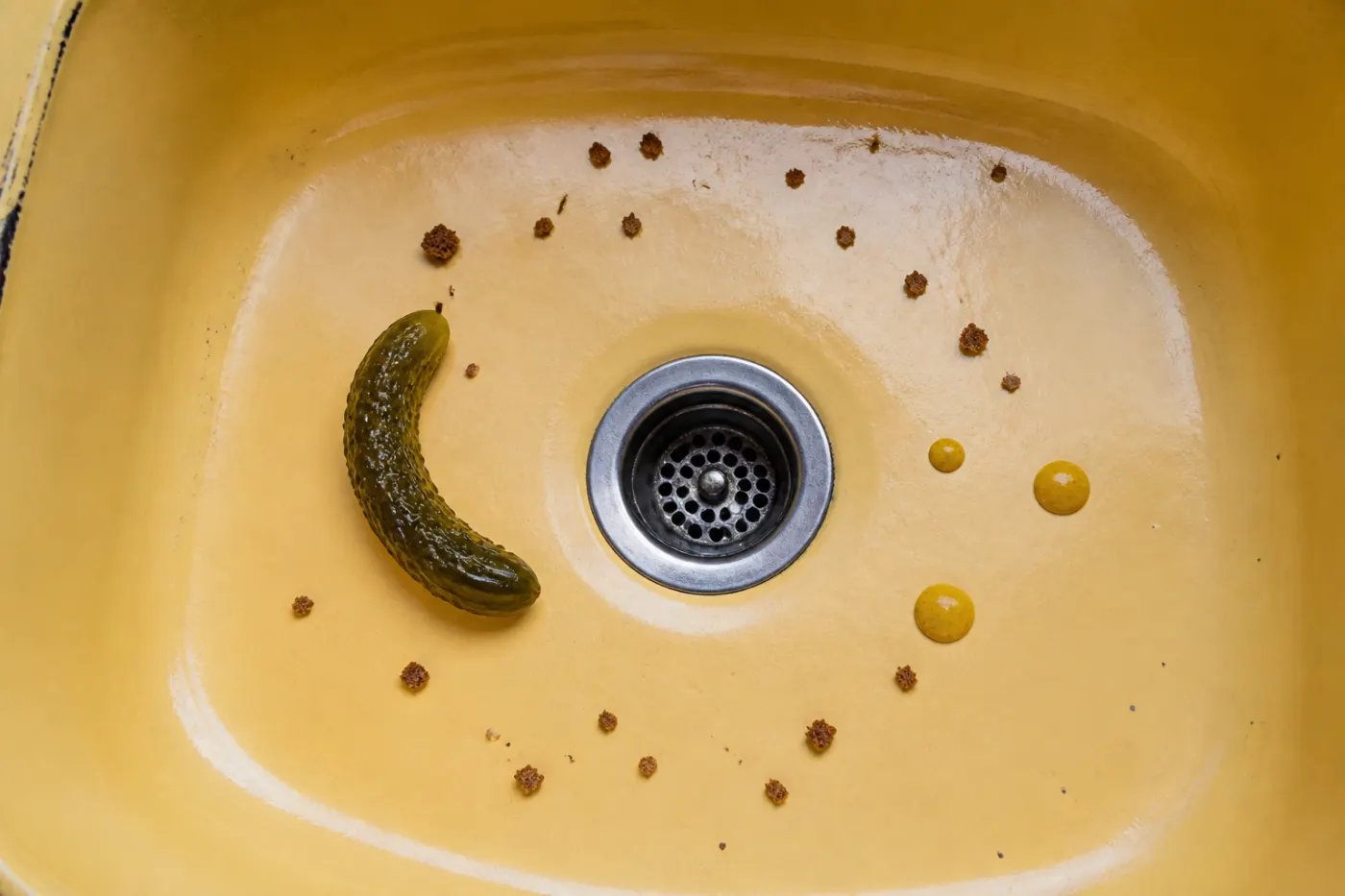 Pickle crescent, mustard droplets and rye crumbs arranged around the drain of a yellow enamel sink