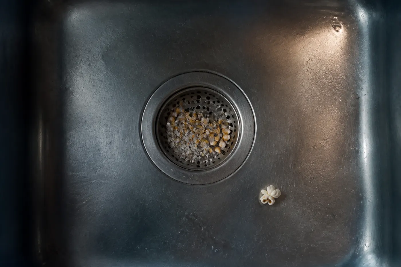 Popcorn hulls and salt caught in a stainless utility sink strainer beside one popped kernel