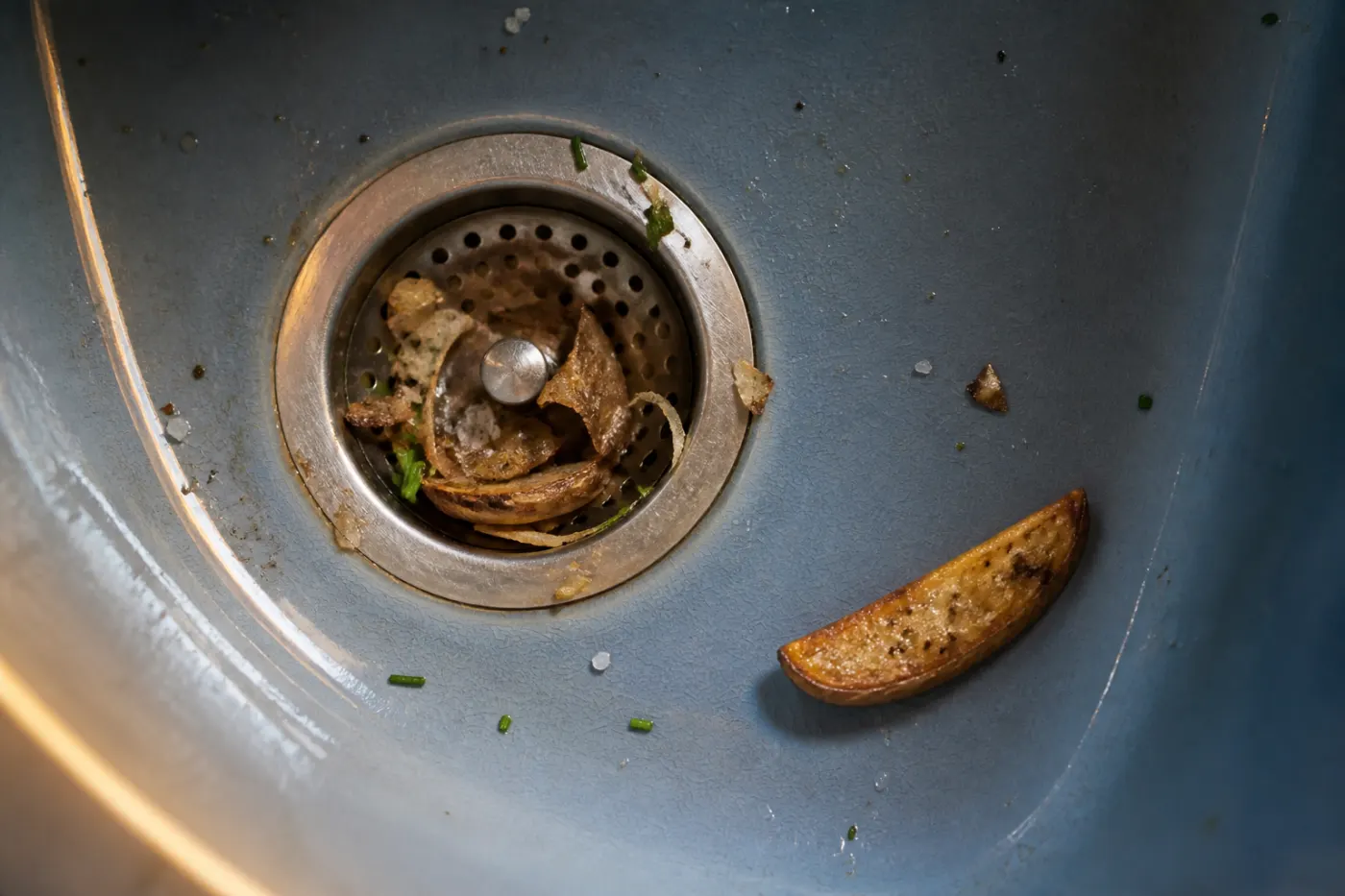 Blue-gray sink with potato skin, chive and salt in the strainer beside a roasted wedge