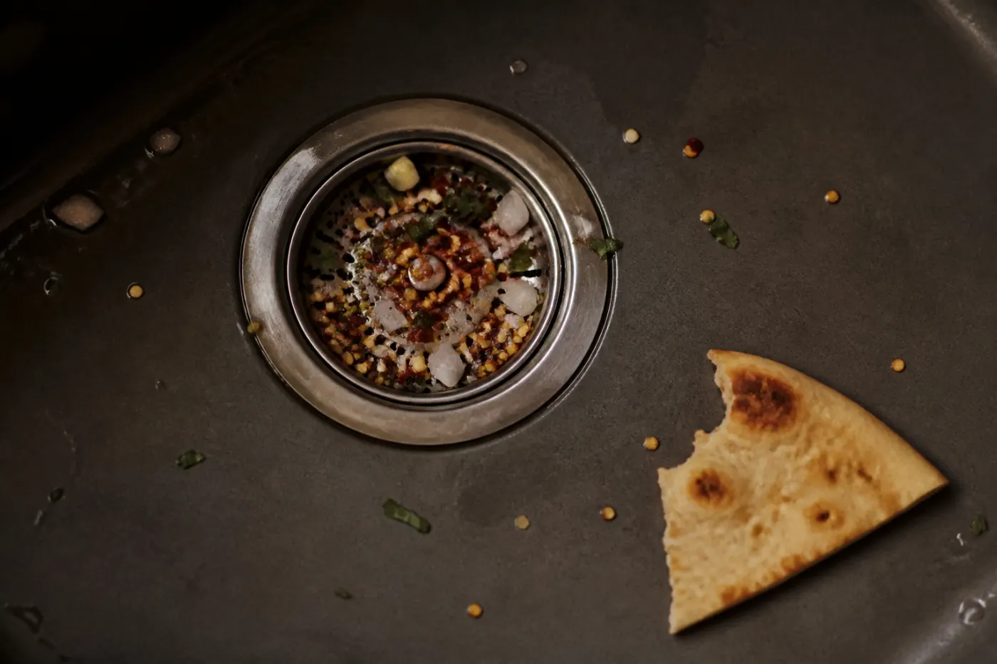 Charcoal sink with pepper seeds, onion and herbs in the strainer beside bitten flatbread
