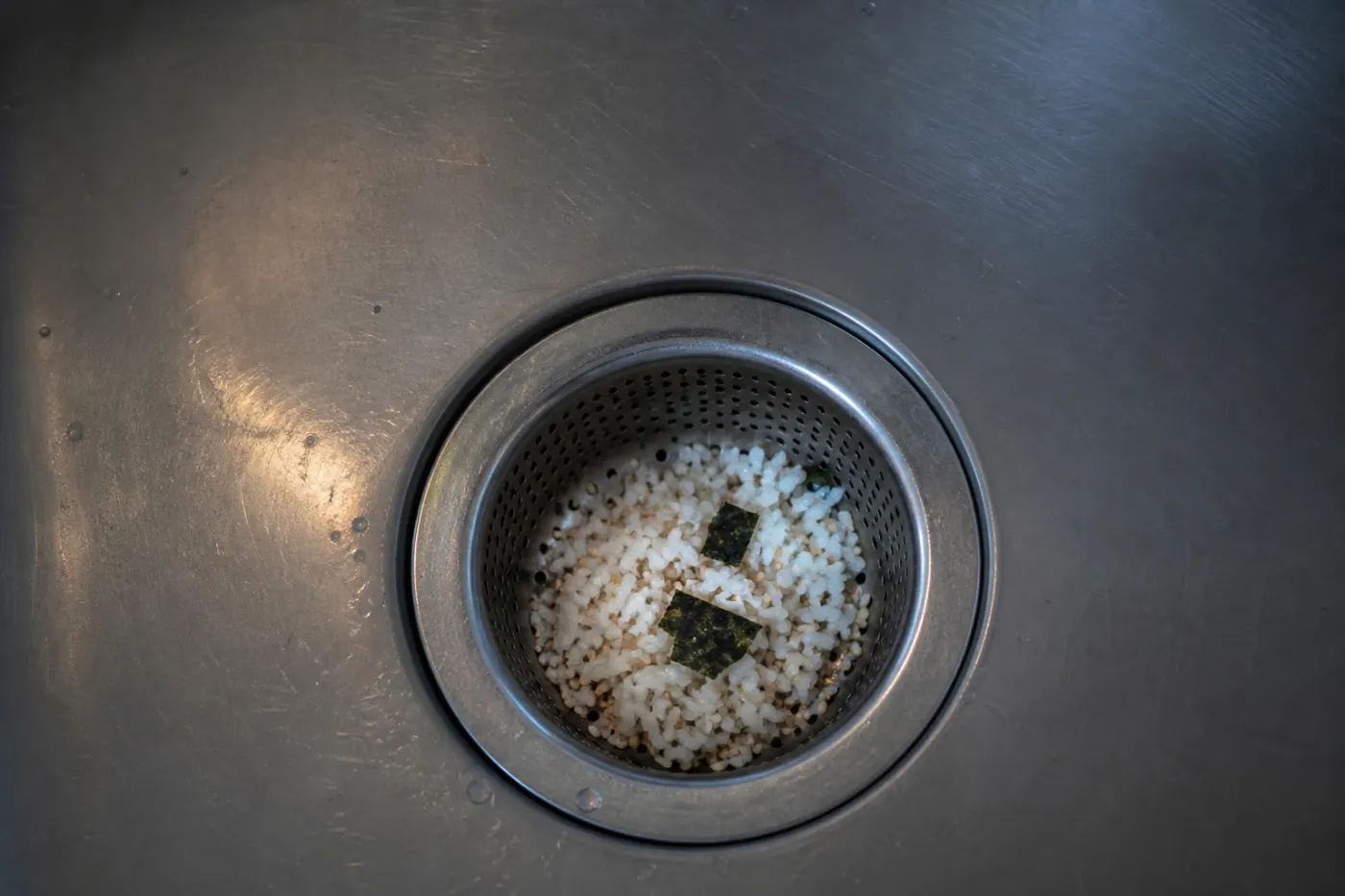 White rice grains, nori squares and sesame caught in a stainless steel sink strainer at night